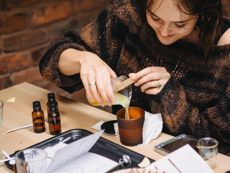 Girl in knitted sweater pouring melted wax into a candle jar